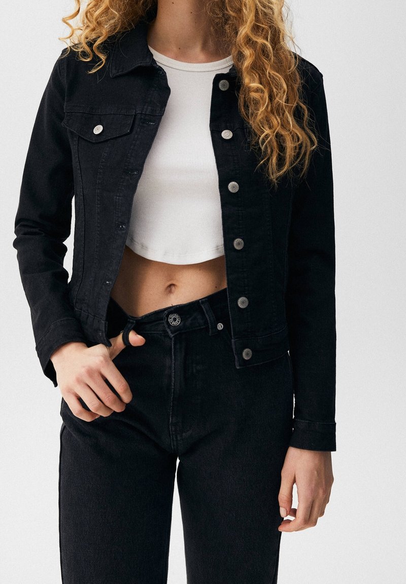 Mujer vestida con chaqueta de mezclilla negra y jeans con la cinturilla desabrochada, top blanco de canalé corto, de pie contra un fondo liso.