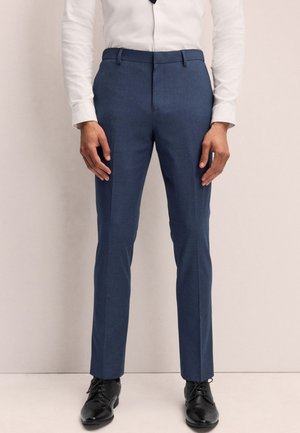 Mężczyzna w dopasowanych niebieskich spodniach garniturowych, białej koszuli i czarnych eleganckich butach stoi na neutralnym tle.