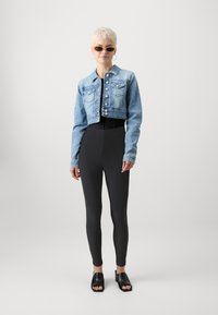 Veste en denim coupée de couleur bleu clair avec des boutons à l'avant, associée à des leggings texturés noirs et des sandales ouvertes noires.