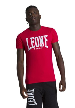 Un homme pose avec un tee-shirt rouge Leone 1947 et un short noir sur fond blanc.