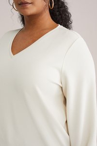 Haut à manches longues et col en V en tissu texturé crème, avec une coupe décontractée et des détails de couture visibles le long des coutures.