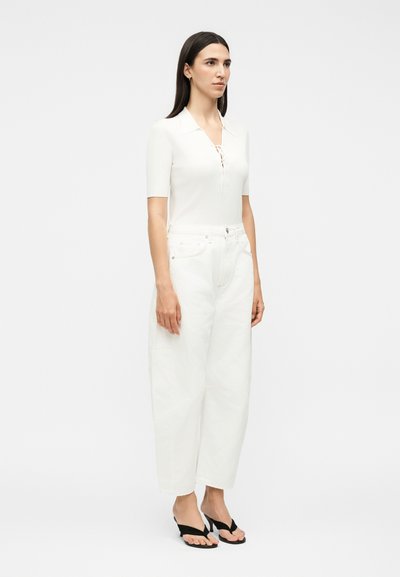 Γυναίκα με ίσια σκουρόχρωμα μαλλιά, ντυμένη με λευκό κοντομάνικο μπλουζάκι, λευκό φαρδύ παντελόνι ψηλόμεσο και μαύρα πέδιλα με ψηλό τακούνι και ανοιχτά δάχτυλα.