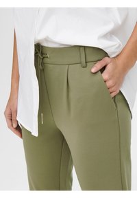 Kalhoty olivově zelené barvy s hladkou texturou, vybavené stahováním v pase a předními sklady. Kombinované s bílou košilí, zobrazující detail bočních kapes.