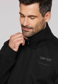 Zwarte zip-up jas met gestructureerde kraag, voorzien van het "CAMP DAVID" logo en aanvullende tekst in grijs. Glad materiaal en fitted ontwerp.
