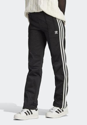 Osoba nosící černé tepláky Adidas s bílými pruhy a bílé tenisky Adidas stojící na bílém pozadí.