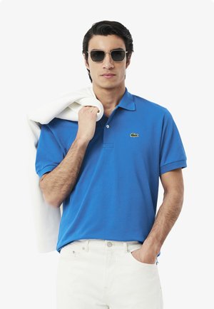 Blå kortärmad polo med klassisk krage och grön broderad logotyp, ihop med vita byxor och en vit tröja över axeln.