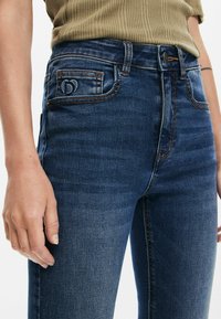 Mörkblå denimjeans med slim fit, detaljerad sömnad och en liten broderad logotyp på bakfickan. Slät textur, klassisk design.