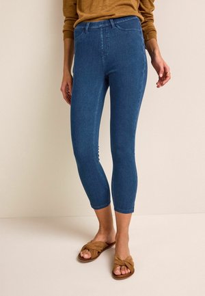 Femme portant un jean skinny court bleu, un haut à manches longues beige et des sandales ouvertes beiges, debout sur un sol et devant un mur unis.