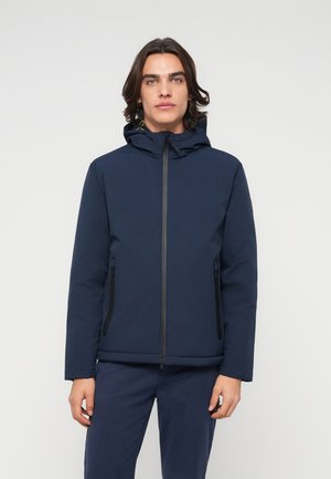 Jeune homme aux cheveux foncés mi-longs portant une veste à capuche bleu marine et un pantalon assorti sur un fond clair uni.