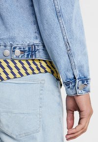 Personne portant une veste en denim bleu clair et un jean, avec une chemise à motif zigzag jaune et marine dépassant au niveau de la taille, le bras détendu au côté.