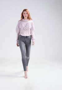 Blusa de color rosa claro con volantes y mangas largas transparentes, combinada con jeans ajustados grises de cintura alta y sandalias de tacón nude.