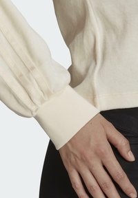 Krämfärgad tyg med texturerade, veckade ärmar. Fitted manschetter och släta ytdetaljer som betonar det mjuka och lätta materialet.