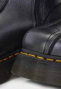 Primer plano de botas de cuero negro con suelas gruesas estriadas y un distintivo pespunte amarillo a lo largo del borde de la suela.