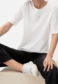 Biała koszulka z krótkim rękawem z małym, okrągłym haftem w czerwono-białe wzory na piersi, połączona z czarnymi dżinsami i białymi trampkami.