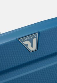 Valigia rigida blu con superficie texturizzata, una patch a logo triangolare con la scritta "RONCATO" e dettagli con chiusura a zip.