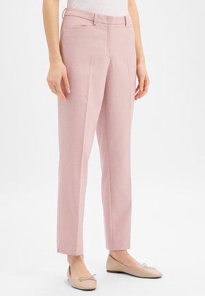 Kobieta ubrana w teksturowane, jasnoróżowe, dopasowane spodnie oraz beżowe baletki, stojąca na tle jednolitego białego tła.