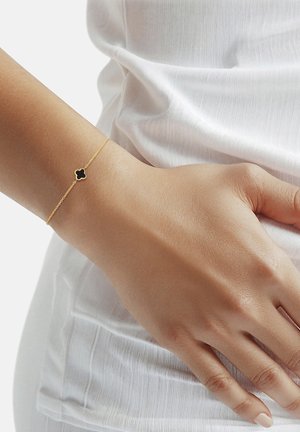 Dünnes goldenes Armband mit einem kleinen schwarzen vierblättrigen Kleeblatt-Anhänger, getragen an einem Handgelenk über einem weißen, strukturierten Stoffärmel.