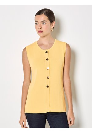 Femme aux cheveux foncés portant un top côtelé jaune sans manches avec cinq gros boutons ronds, associée à un jean foncé, debout contre un mur blanc.