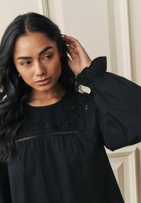 Melna blūze ar izsmalcinātu ziedu izšuvumu apkaklē, ar pūkainām piedurknēm un savilktu ķermeņa tekstūru.