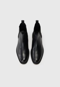 Par de elegantes botas Chelsea de cuero negro con paneles laterales elásticos y lengüetas, vistas desde arriba sobre un fondo blanco.