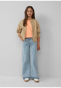 Beige bomberjack, lichtoranje t-shirt, wijdvallende lichtblauwe jeans, witte schoenen en steil bruin haar. Eene eenvoudige achtergrond.