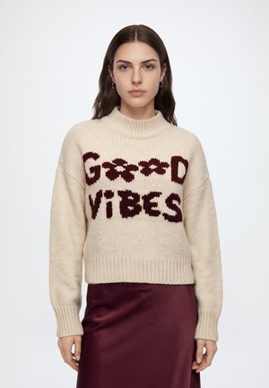 Femme aux longs cheveux foncés portant un pull crème avec "GOOD VIBES" en lettres rouge foncé et une jupe en satin rouge foncé sur fond blanc.