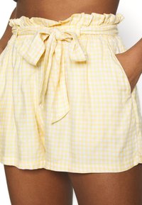 Pohled z pasu dolů na žluté a bílé kostkované šortky z papírového pytle s uvázaným látkovým páskem a bočními kapsami, které má na sobě osoba.