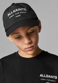 Svart keps med vit broderad text "ALLSAINTS" och ytterligare designelement; bärs med en svart tröja som har liknande branding.