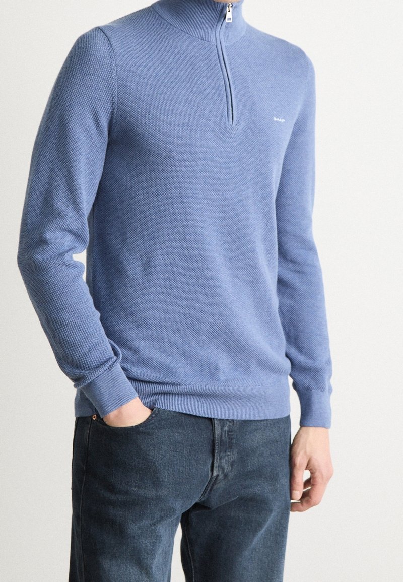 Himmelblaues Zip-Pullover aus strukturiertem Stoff, mit langen Ärmeln und gerippten Bündchen sowie Saum. Kombiniert mit dunkelblauen Jeans.