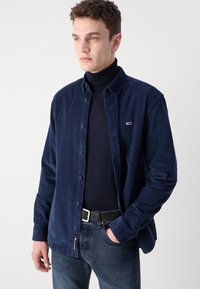 Camicia in velluto a coste blu navy con bottoni e maniche lunghe, indossata sopra un dolcevita nero, presenta un piccolo logo ed è abbinata a jeans scuri e una cintura.