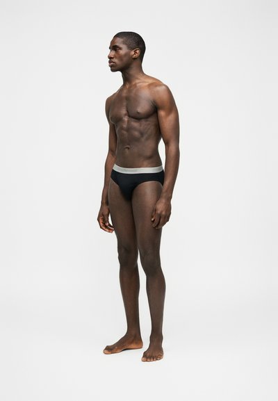 Άνδρας μοντέλο όρθιος ξυπόλητος, φοράει μαύρα σλιπ με ανοιχτόχρωμη μέση, σε απλό ανοιχτό φόντο, στραμμένος προς τα αριστερά.