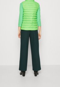 Gilet imbottito verde chiaro sopra una maglietta turchese a maniche lunghe, abbinato a pantaloni larghi verde scuro. Il tessuto è liscio con una finitura lucida.