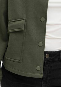 Olive zaļš džemperjaka ar teksturētu apdari, ar sānu kabatu un apaļām pogām, kombinēta ar melnajiem džinsiem.