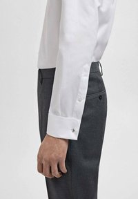 Camisa blanca de manga larga con puños puntiagudos, que presenta un gemelo de plata, combinada con un pantalón oscuro gris en un corte ajustado.