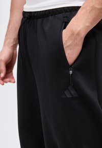 Gros plan d’une personne portant un pantalon Adidas noir avec une poche zippée, main partiellement dans la poche, et une chemise blanche visible.