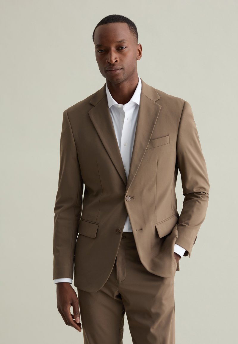 Blazer marrone con pantaloni abbinati, caratterizzato da una vestibilità slim, rever a notche e chiusura con un solo bottone, abbinato a una camicia bianca con colletto.