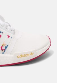 Adidas sneaker featuring a white fabric upper with floral accents, textured finish, gold logo, and pink outsole.