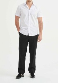 Chemise blanche à manches courtes avec boutons, associée à un pantalon noir et des chaussures noires. La chemise présente un col classique et des lignes épurées.