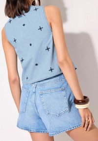 Žena v světle modrém tílku bez rukávů s černými vyšívanými kříži a odpovídajících džínových šortkách, doplněná masivními náramky.