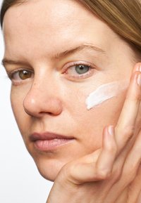 Hydratant appliqué sur une joue ; texture crémeuse et blanche. La peau est lisse avec de subtiles taches de rousseur. Les cheveux sont châtain clair et coiffés raides.