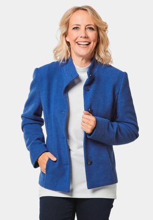 Blauwe fleece jas met hoge kraag, twee knopen en zakken. Glad textuur en getailleerde pasvorm, gedragen over een wit topje en donkergekleurde broek.