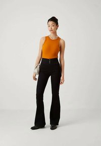 Jeune femme portant un haut sans manches orange, un pantalon noir évasé taille haute, des chaussures noires, tenant un sac à main beige, se tenant sur un fond uni.