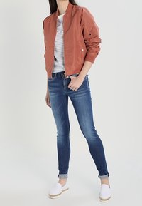 Rostfärgad bomberjacka, randig vit t-shirt, mörkblå skinny jeans, vita slip-on skor med jute-sulor. Slät textur, modern design.