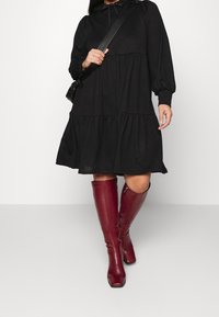 En person klädd i en svart, lager-på-lager klänning och knälånga bordeauxfärgade stövlar håller en svart väska och står mot en neutral bakgrund.