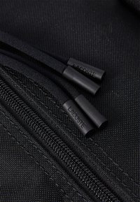 Detalle de cremallera negra con tiradores de goma con la marca "LACOSTE", sobre un tejido texturizado, destacando la pista de cremallera metálica suave.