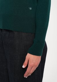 Tmavě zelený svetr s ribovanou strukturou a přiléhavými manžetami. Výrazný detail loga ve světle modré na lemu. Spárováno s tmavými džíny.
