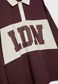 Burgundy polo-shirt med hvide detaljer, der har et stort "LDN" grafik. Lavet af bomuld, har den en klassisk krave og knaplukning.