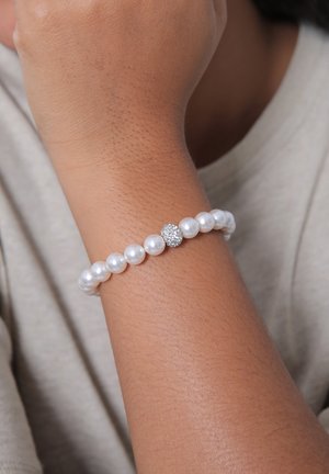 Perlenarmband mit abwechselnd glänzenden weißen Perlen und einer zentralen runden silberfarbenen Perle mit Glitzer, die auf einem gebräunten Handgelenk ruht.