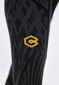 Κοντινό πλάνο μαύρων κολαν με σκούρα αφηρημένα μοτίβα και μικρό κίτρινο εξαγωνικό λογότυπο στο μηρό.