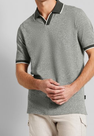 Mann mit grauem Kurzarm-Poloshirt mit schwarz-weißem Kragen und Bündchen, die Hände vor dem Körper verschränkt, beige Hose sichtbar.